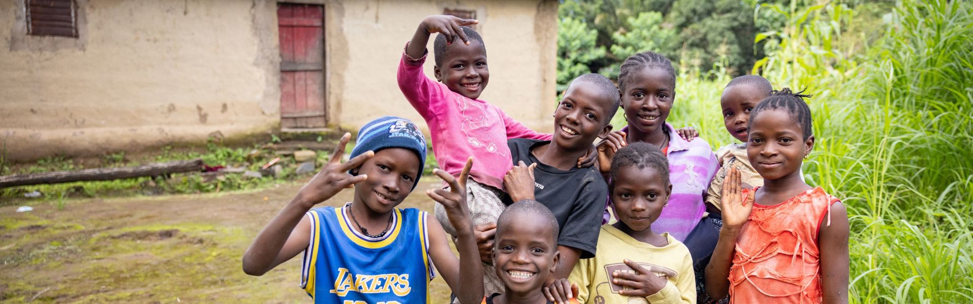 A group of children in Guinea stand outside of a hut, smiling at the camera.