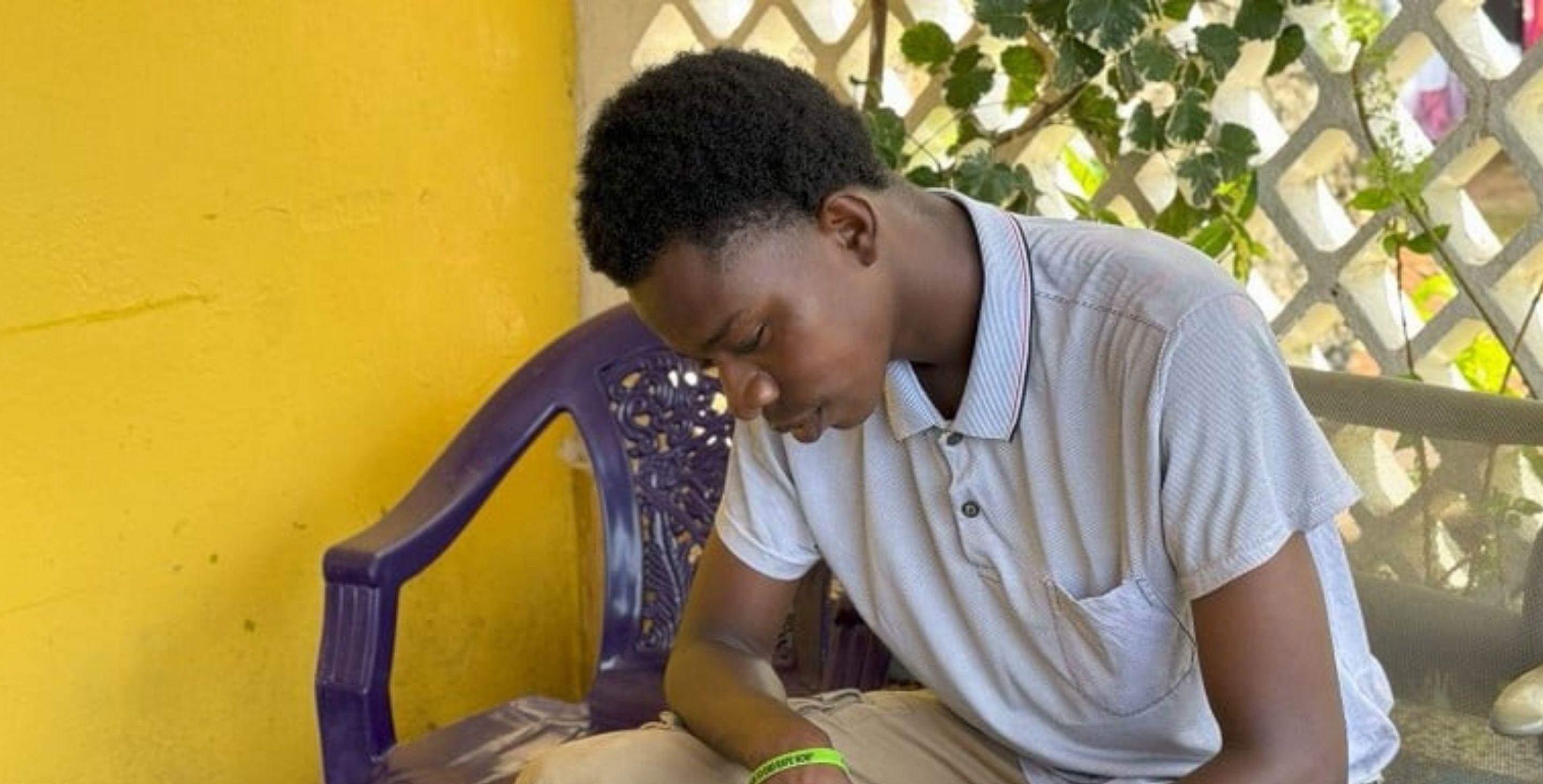 A teenage boy in Sierra Leone sits on a porch outdoors, leaning over something he is writing.