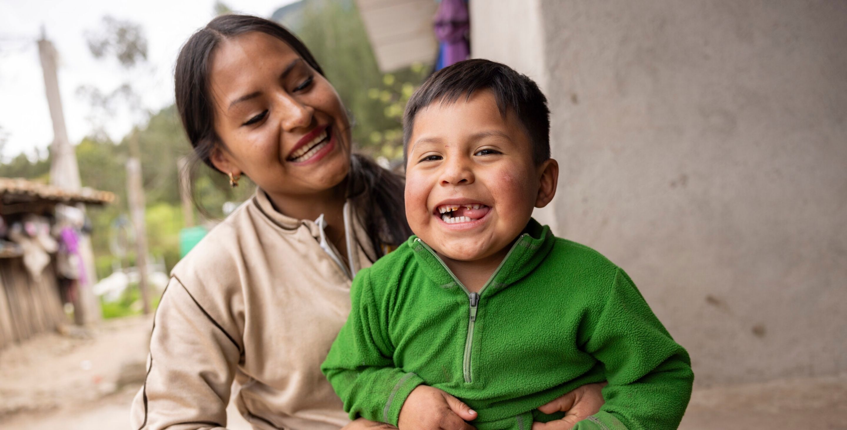 A mother sits on the ground holding her toddler in her arms, who is smiling at the camera.