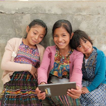 Three girls in Guatemala sit against a wall, looking at a tablet and smiling.