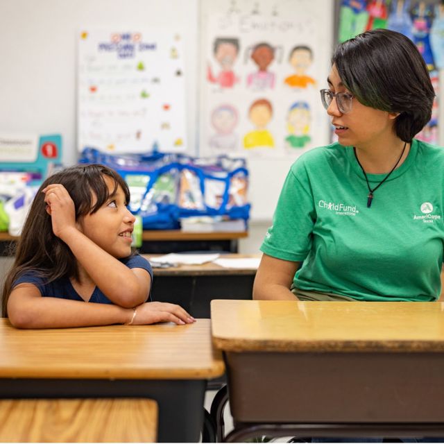 A young woman in a ChildFund shirt and a little girl sit at a desk in an American elementary school, talking.