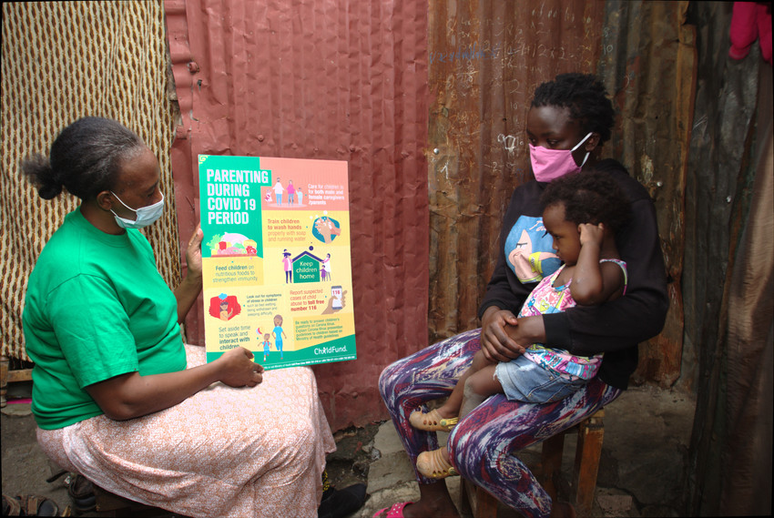 Woman wearing a green ChildFund shirt and a mask speaks to a mother and her young child while holding a poster labeled “Parenting During COVID-19 Period.”