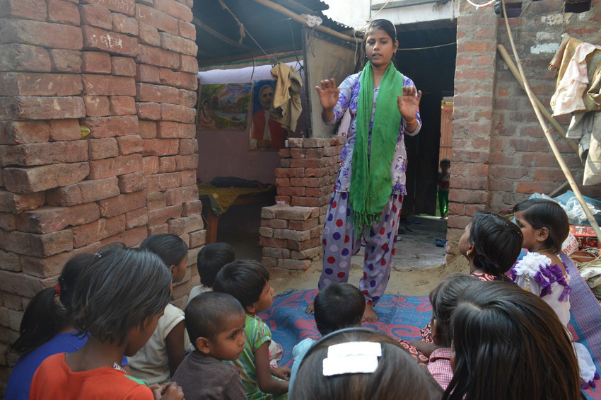 A teenage girl stands up while speaking to a group of seated children in a courtyard in India.
