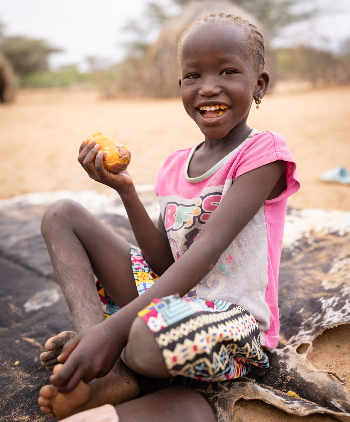 A little girl in Kenya sits on a mat in a dusty landscape, eating a sweet potato and smiling.