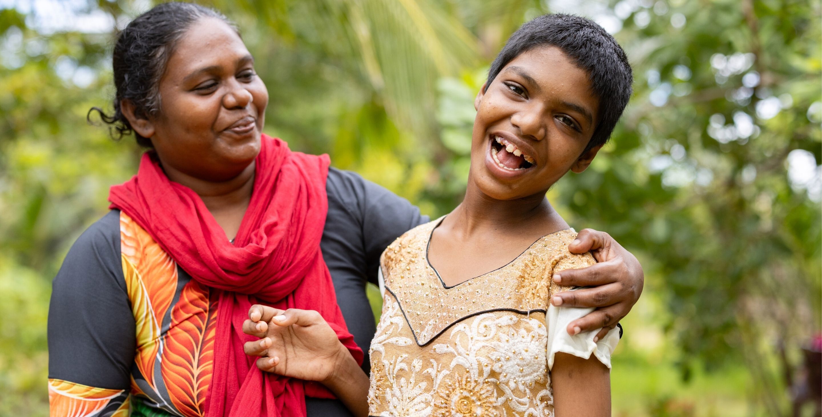 A mom stands with her teenage daughter, with her arm around her, smiling at her as the daughter smiles widely at the camera.