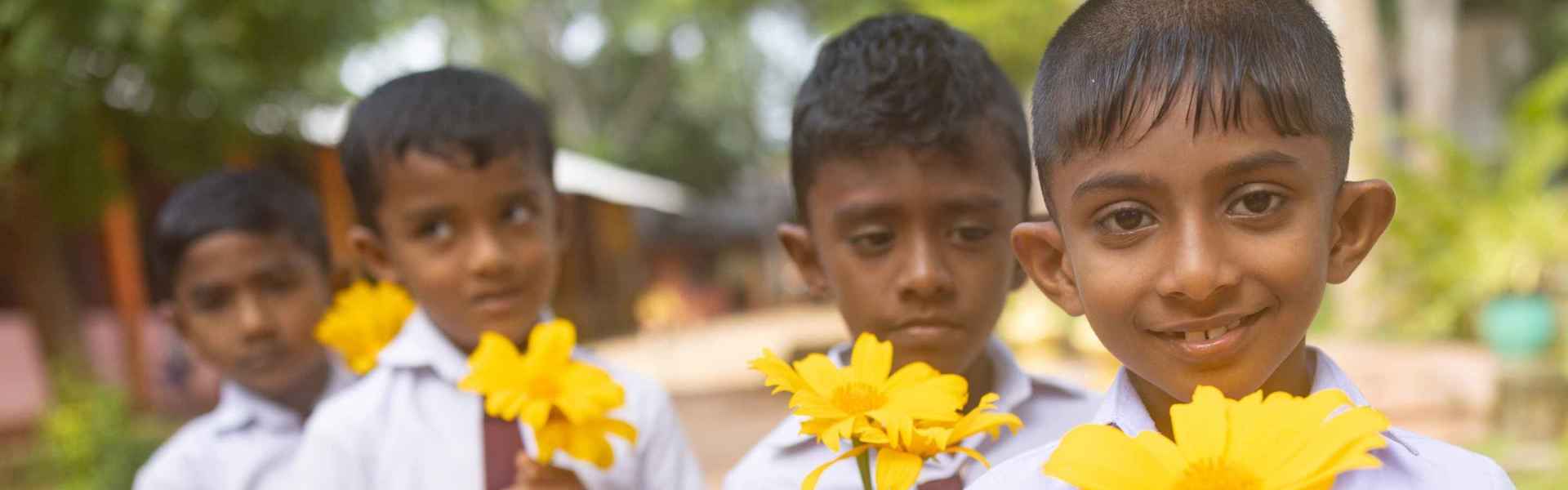 four primary school boys in school uniform holding yellow flowers; boy in foreground is smiling