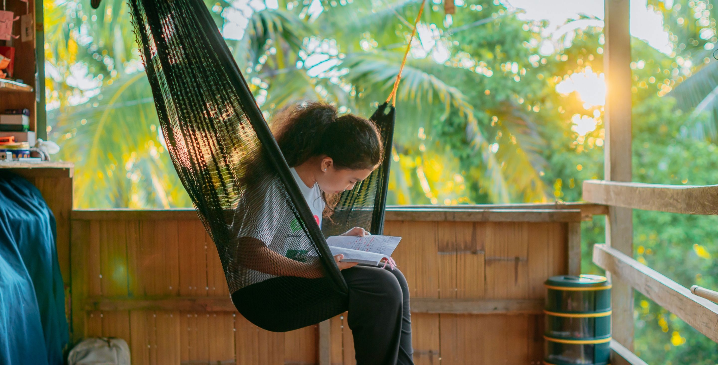 A girl in the Philippines sits in a hammock in her house, studying, as the sun shines behind her.
