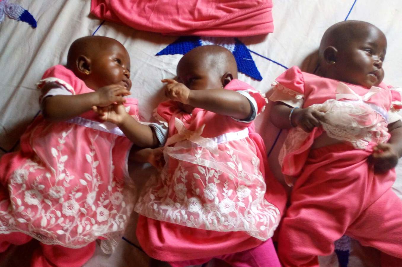 Three infant girls in Guinea lay on a mattress wearing frilly pink dresses.