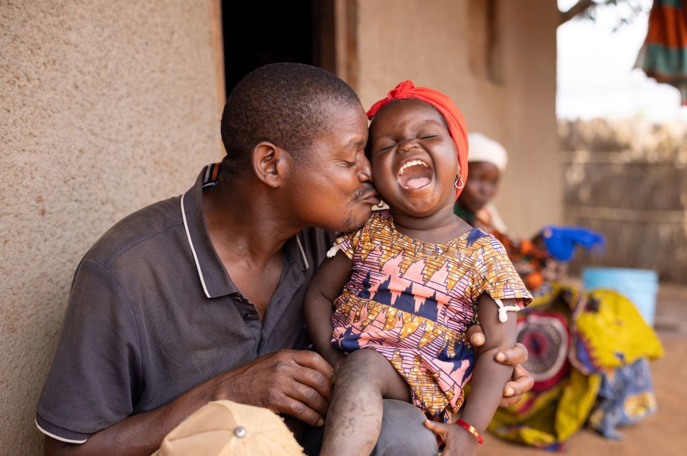 A father and his young daughter sit outside a home in Mozambique. The father is kissing his daughter's cheek and the little girl is throwing her head back in a laugh.
