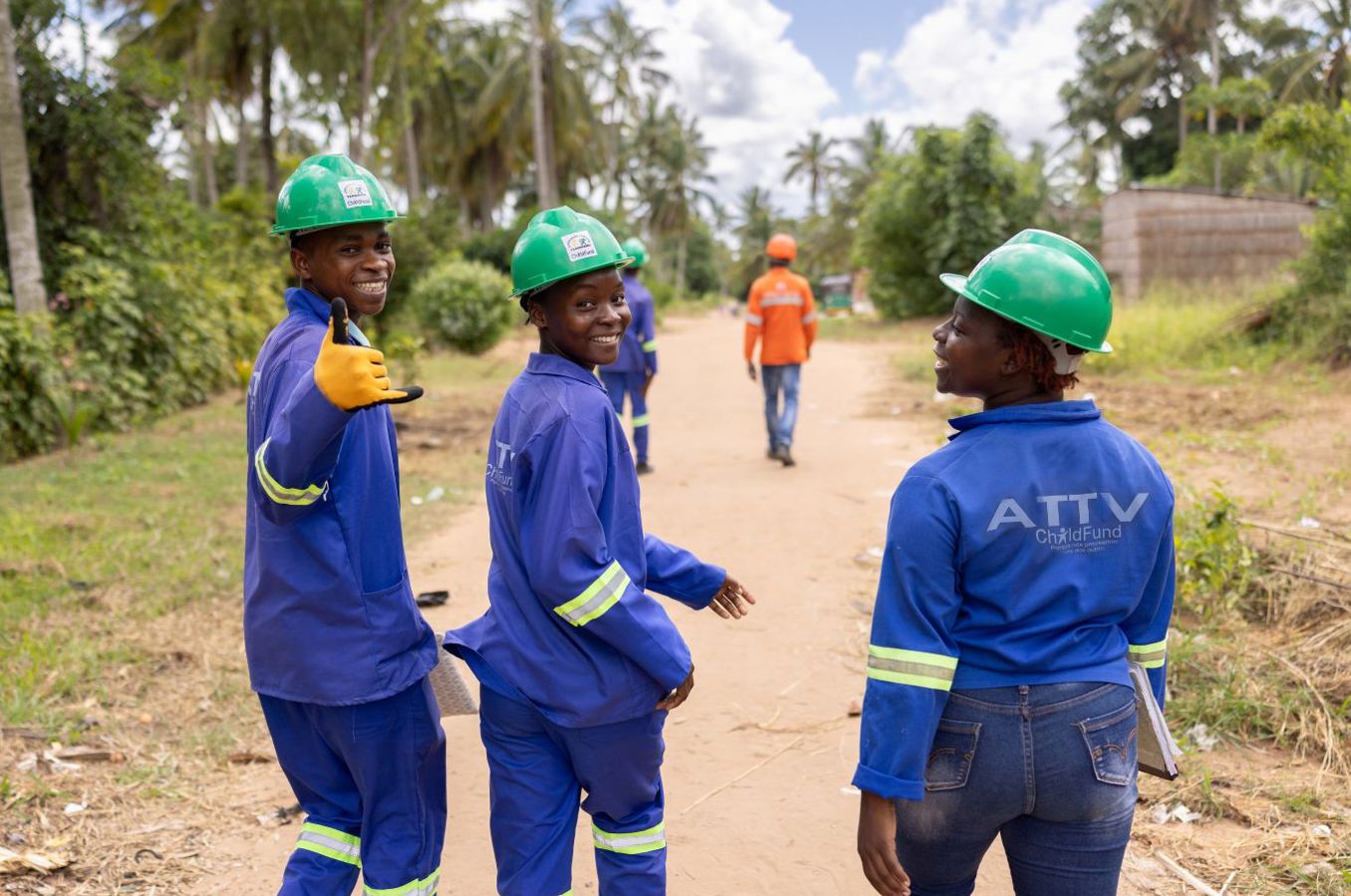 Three youth walk down a dusty road in Mozambique wearing electrician uniforms and hard hats, turning around and waving at the camera. Behind them, their supervisors are leading the way down the road.