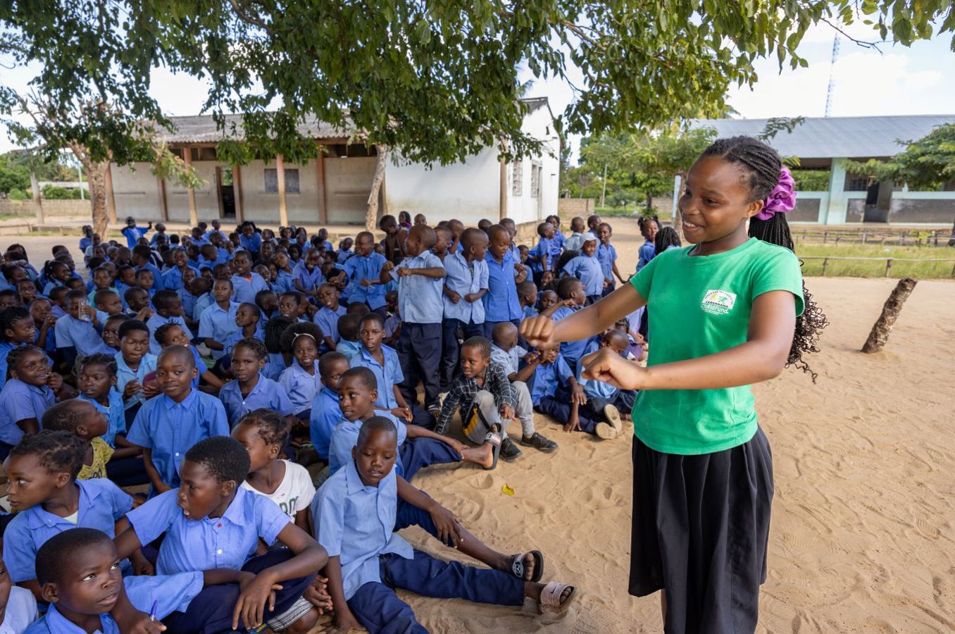 A girl wearing a green shirt teaches a class outside under a tree in Mozambique.