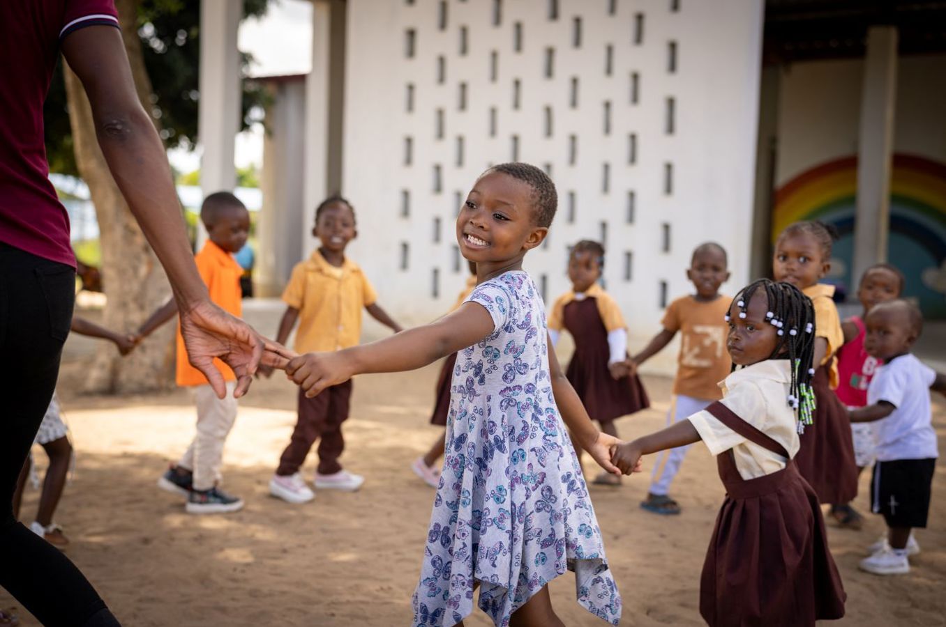 A group of children hold hands in a circle at an ECD center in Mozambique. The shot centers on a little girl wearing a blue dress and smiling happily. 