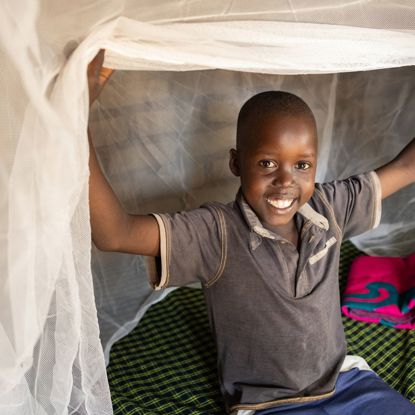 A little boy sits on a bed under a mosquito net, lifting the net up and smiling at the camera.