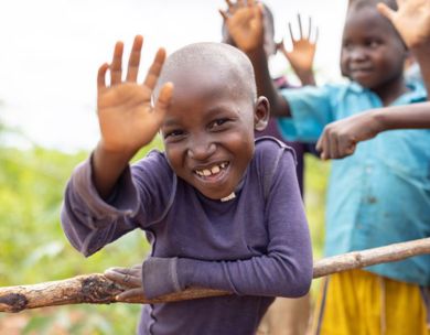 A smiling young boy in a purple shirt waves at the camera while leaning on a wooden fence, joined by other cheering children in a rural Mozambican village.