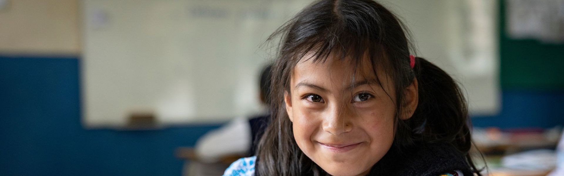 A little girl in Ecuador dressed in traditional clothing sits at a desk writing, smiling at the camera.