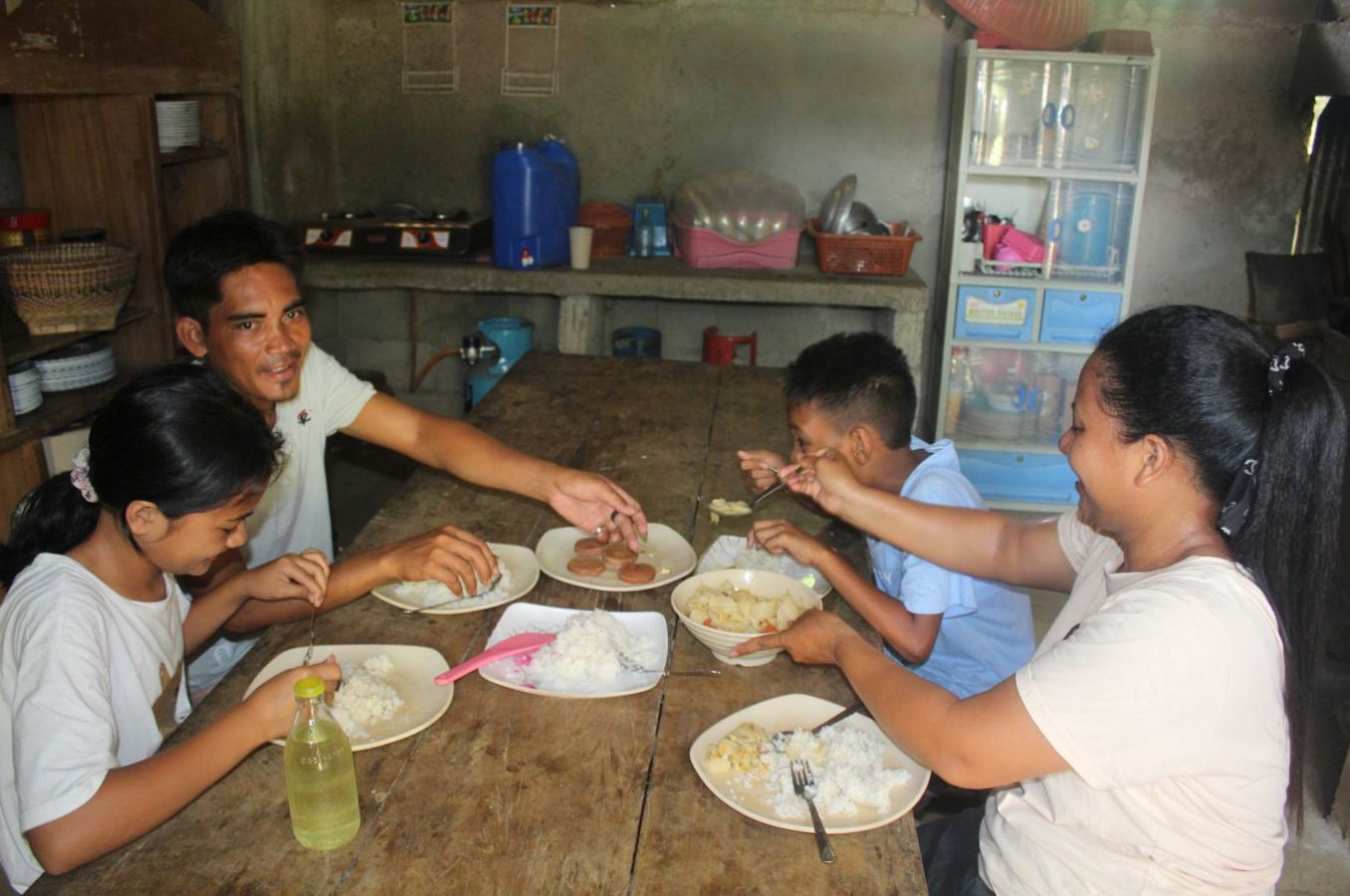A family sits around a wooden table in a home in the Philippines, smiling as they pass food around.