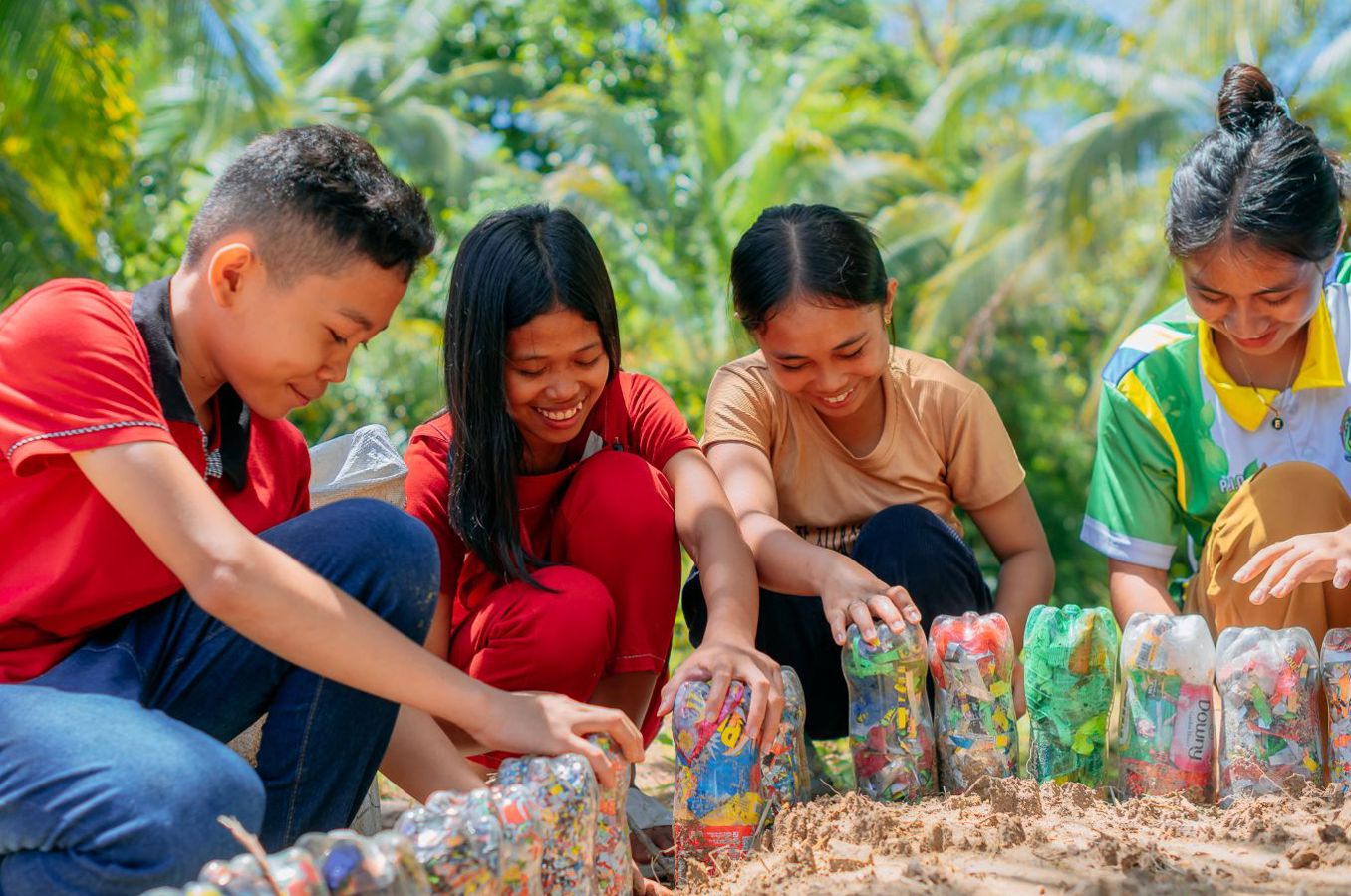 A group of teenagers gather around a community garden featuring a perimeter of reused plastic bottles, smiling as they work in the dirt.