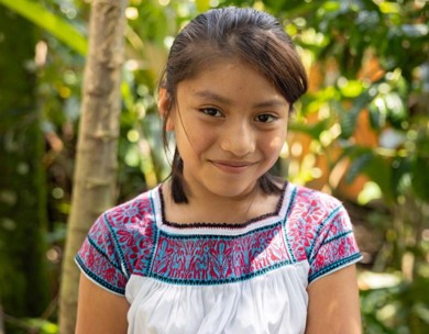 A young girl with dark hair tied back, smiling warmly while wearing a white dress with blue and red geometric embroidery, standing in a sunlit forest.