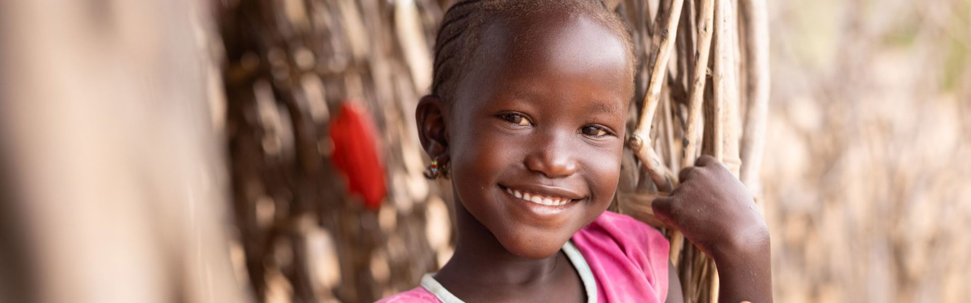 A little girl wearing a pink shirt stands in the doorway of a thatched hut, smiling.