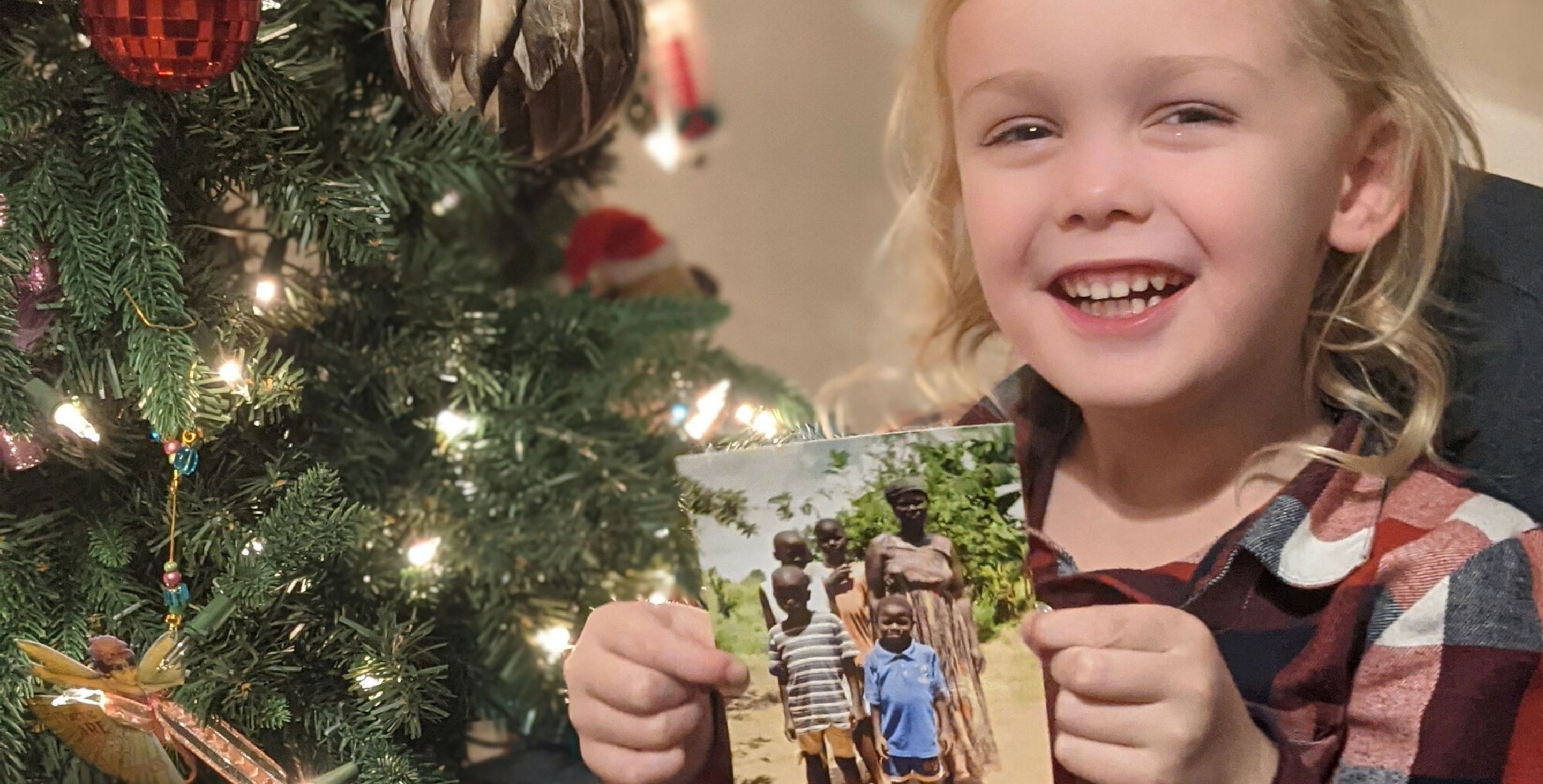 A little boy seated in front of a Christmas tree holds up a photo of his sponsored family, smiling.