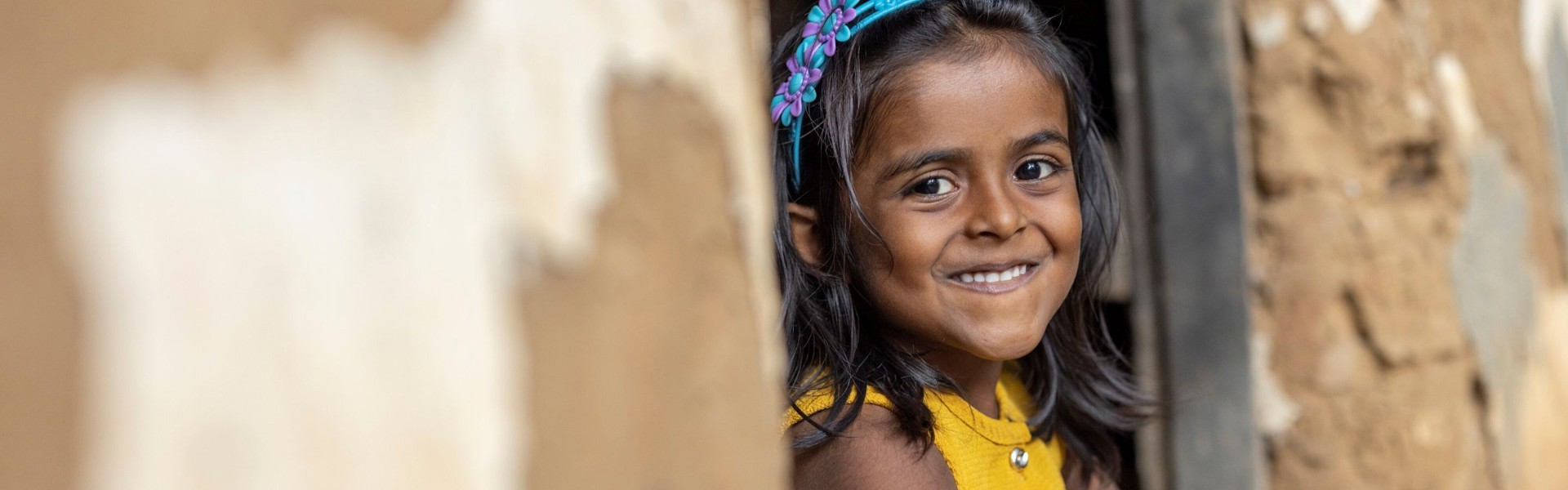 A little girl sits in the doorway of a mud home in Sri Lanka, smiling at the camera.
