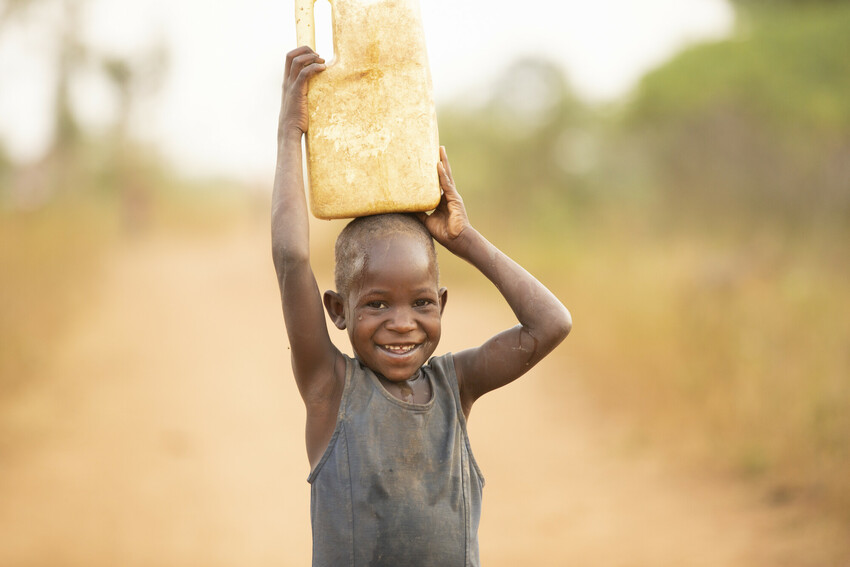 A girl with a bucket of water stands smiling at camera in Uganda.