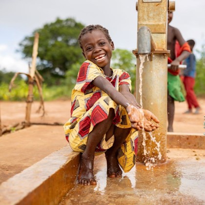A young girl in Mozambique wearing colorful traditional dress washes her hands at a water well.