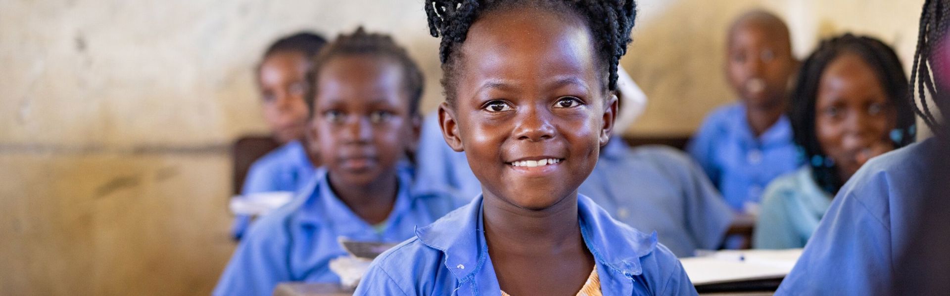 A young girl wearing a blue school uniform sits at a desk in her classroom in Mozambique, smiling.