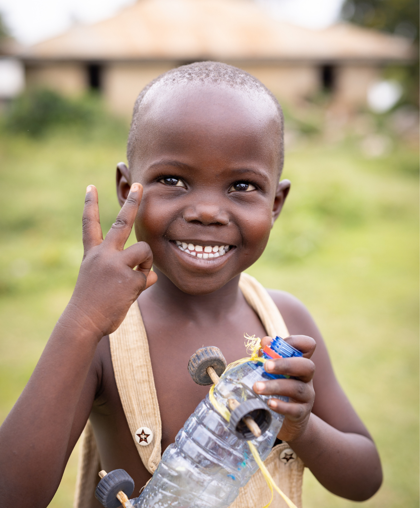 A little girl in Kenya wearing overalls holds up a toy car she made out of a plastic bottle and bottle caps.