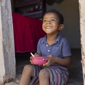 A little boy sits in a doorway in Brazil, eating out of a pink bowl.