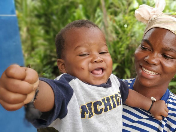 A happy baby in a navy and gray t-shirt reaches out while being held by their smiling mother, who wears a striped shirt and headscarf, with greenery in the background.
