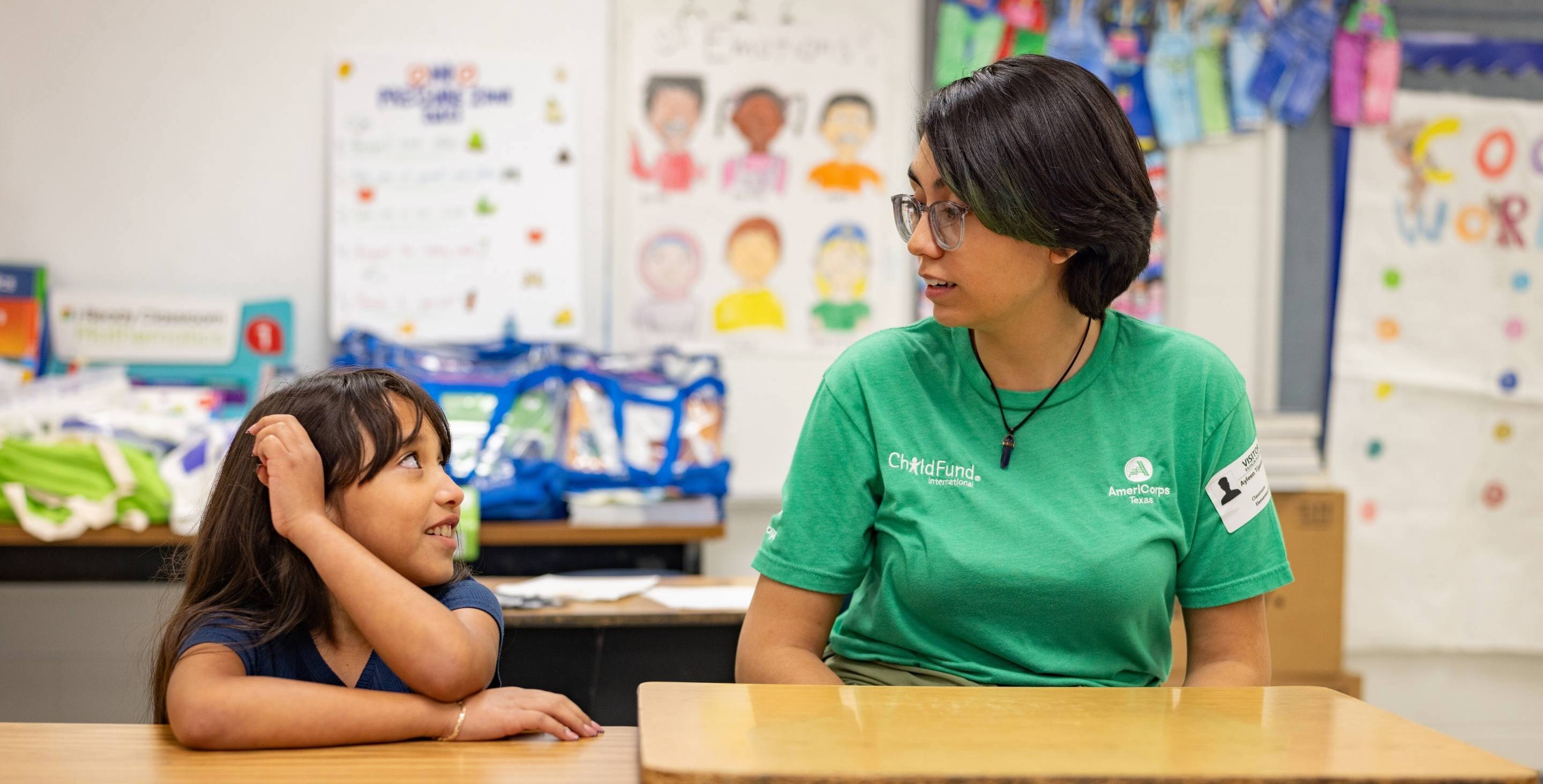 A young woman and little girl sit next to each other at desks in an elementary school, looking at each other and talking.