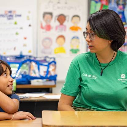 A young woman and little girl sit next to each other at desks in an elementary school, looking at each other and talking.