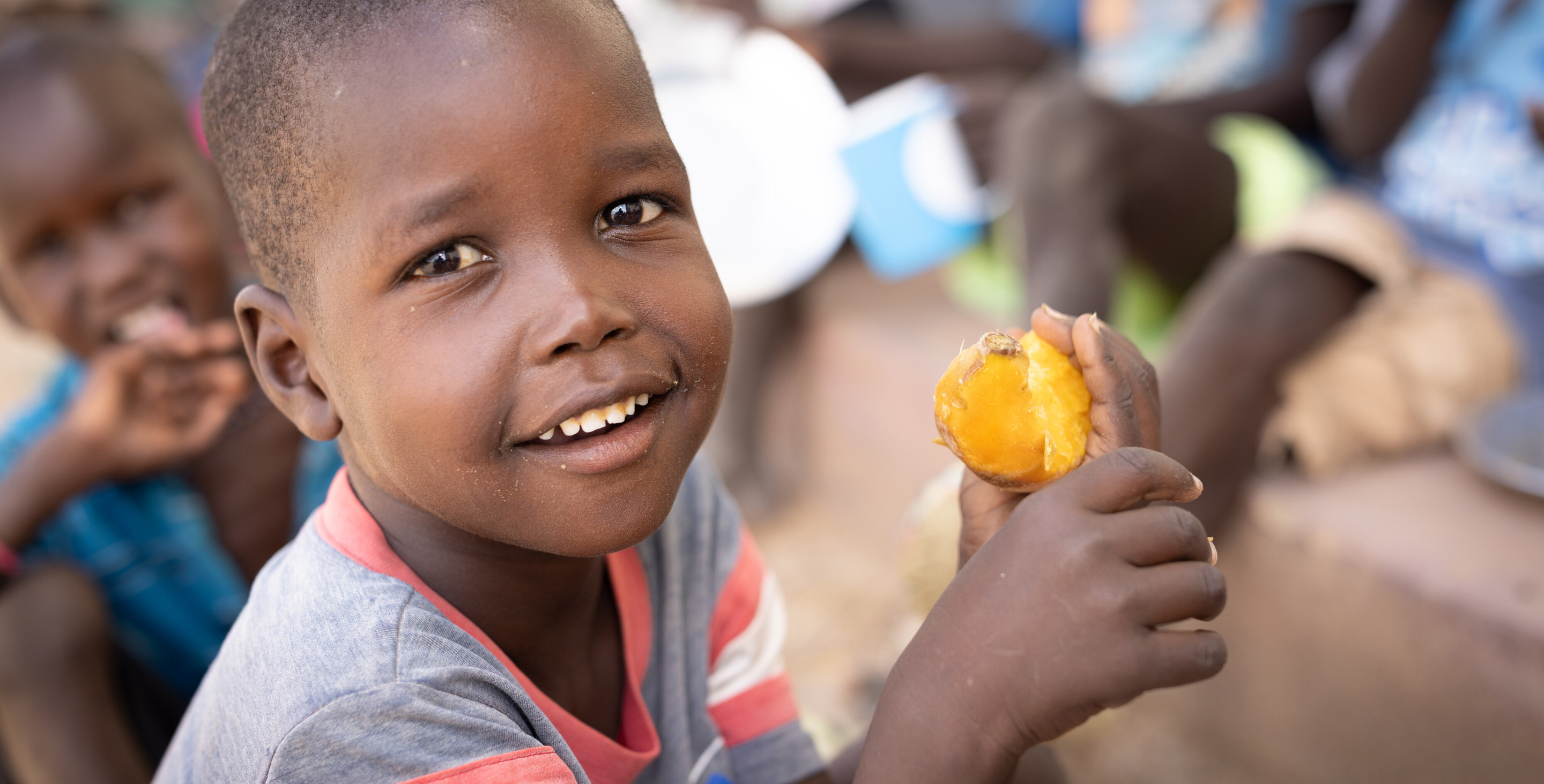 A boy eats a sweet potato in Kenya while smiling at the camera.