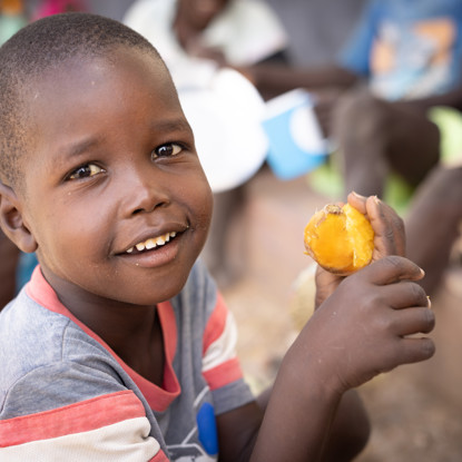 A boy eats a sweet potato in Kenya while smiling at the camera.