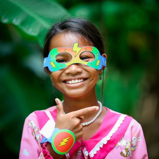 A girl in Cambodia wearing a superhero mask smiles at the camera, pointing to the text on the right.