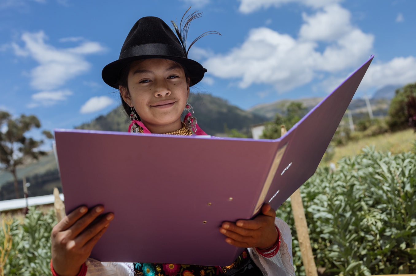 A girl in Ecuador wearing a traditional dress and hat sits in a field, reading a book.