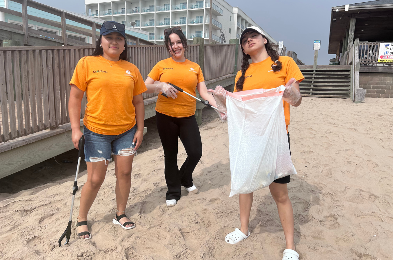 Three teenage girls in orange shirts stand on a beach smiling, carrying trash pickers and trash bags.