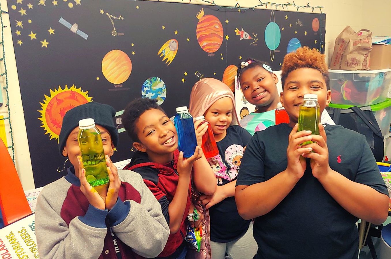 A group of children hold up calming bottles they made as an art project, smiling in a U.S. classroom.
