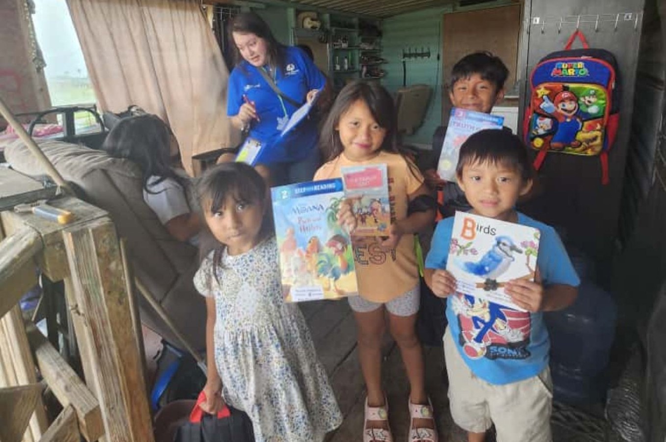 A group of children stands in a small room in a home, holding books and smiling. There is a ChildFund staff member smiling in the background.