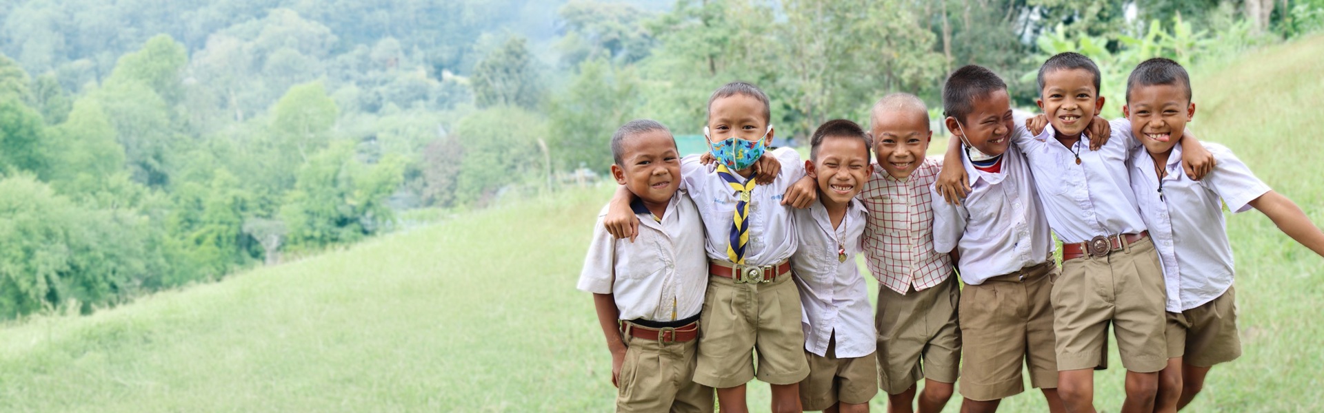 A group of school-aged boys stands in a line in a field in Thailand, laughing with their arms around each other.