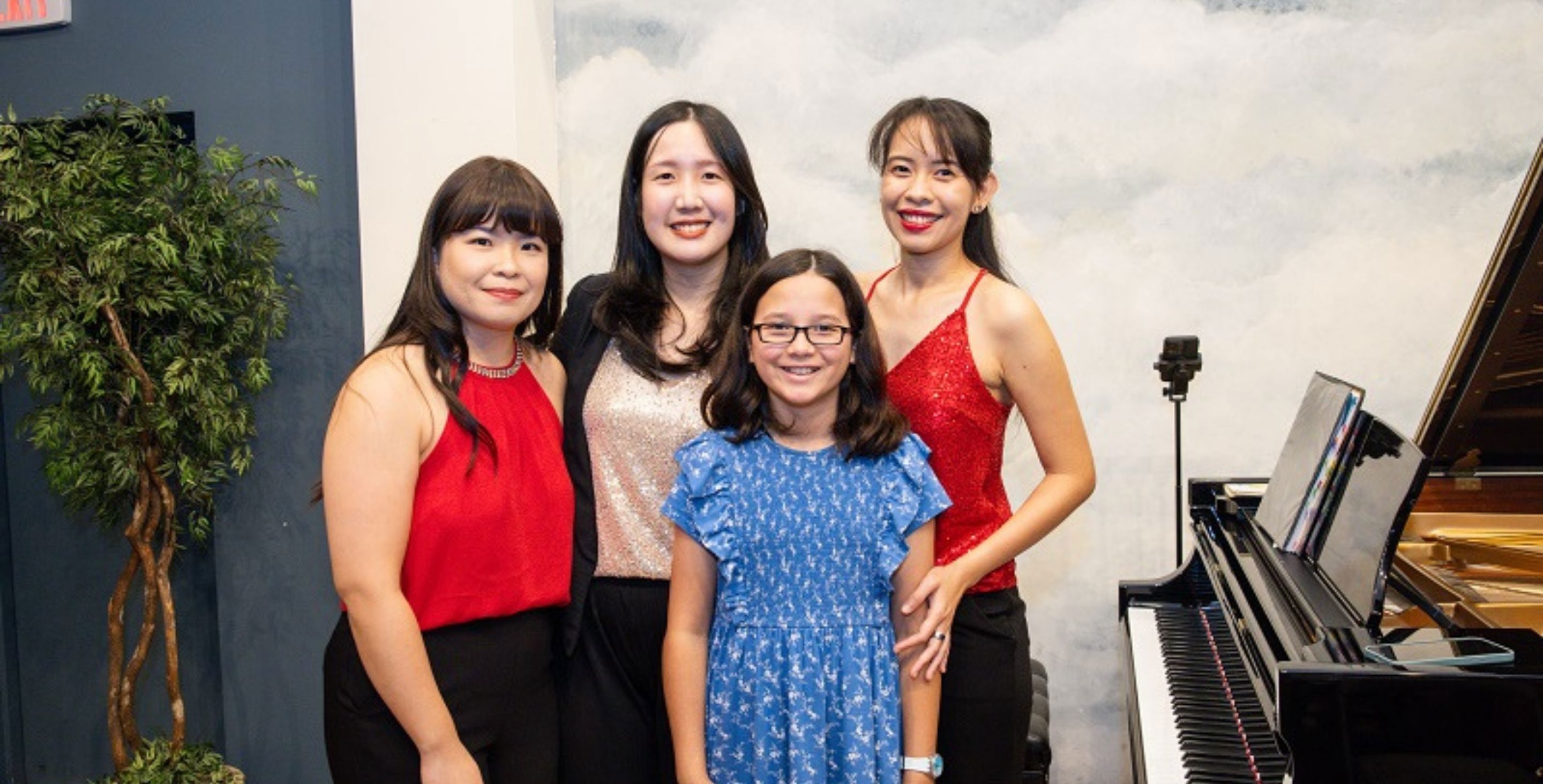 Three young women stand around a piano with a female student, dressed well, apparently for a piano recital.
