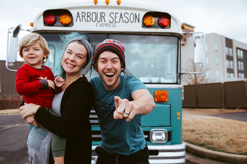 A man, woman and small child stand in front of a blue school bus that reads 'Arbour Season'