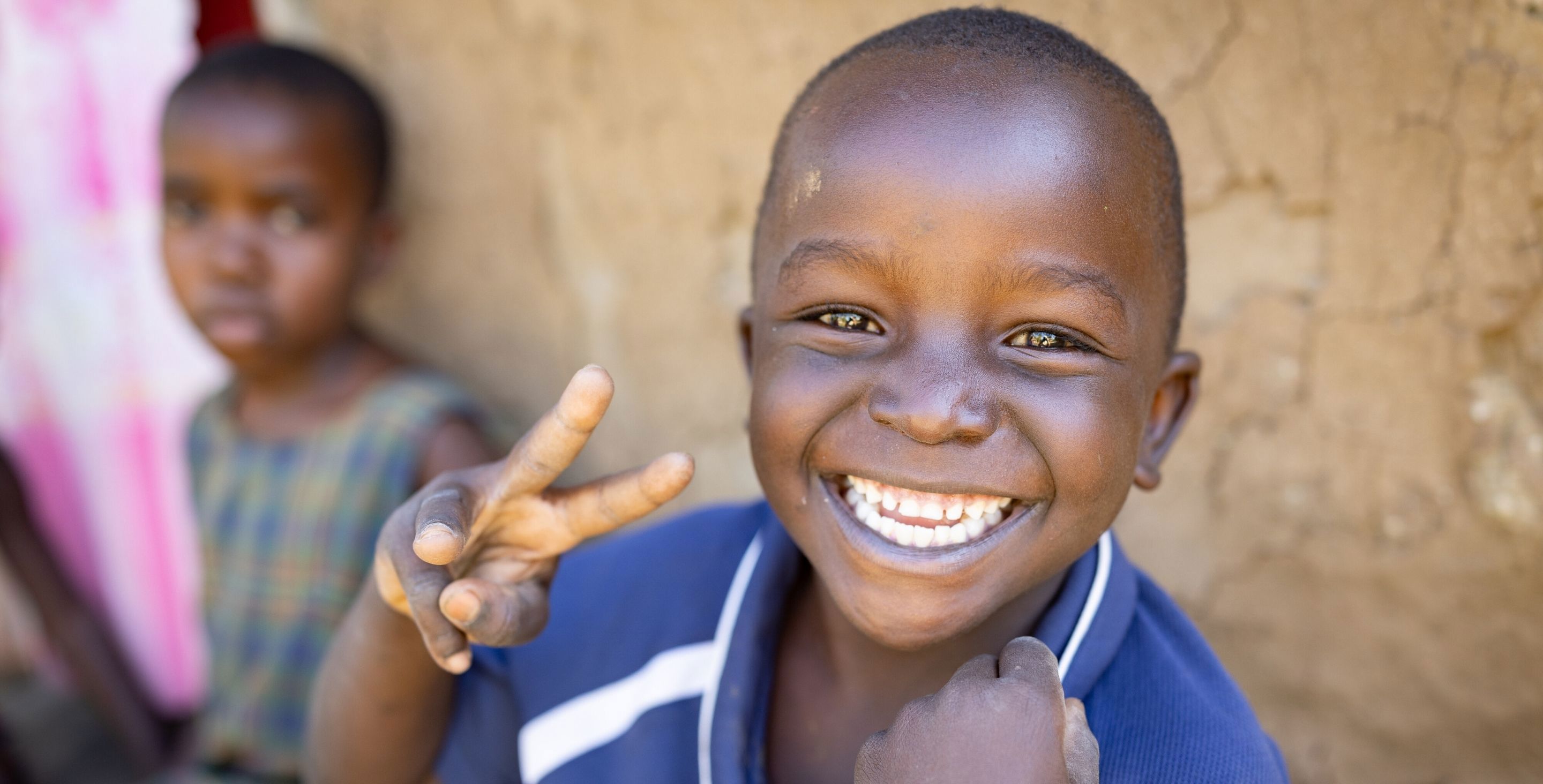 A little boy sitting outdoors in front of an earthen home flashes a peace sign.