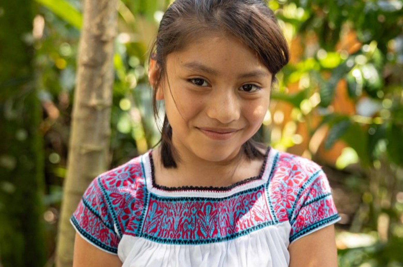 A young girl with dark hair tied back, smiling warmly while wearing a white dress with blue and red geometric embroidery, standing in a sunlit forest.