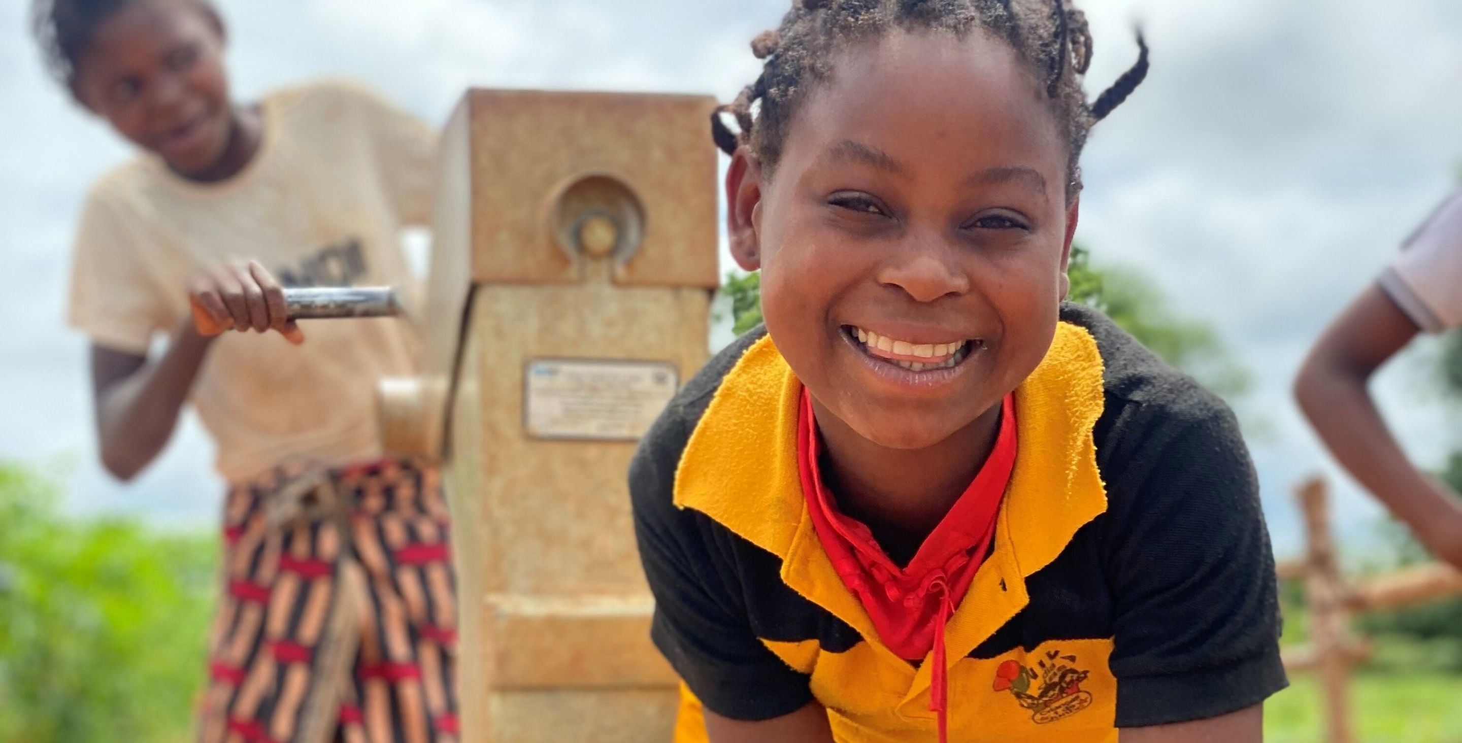 A little girl in Mozambique smiles as she collects water from a borehole into a large bucket.