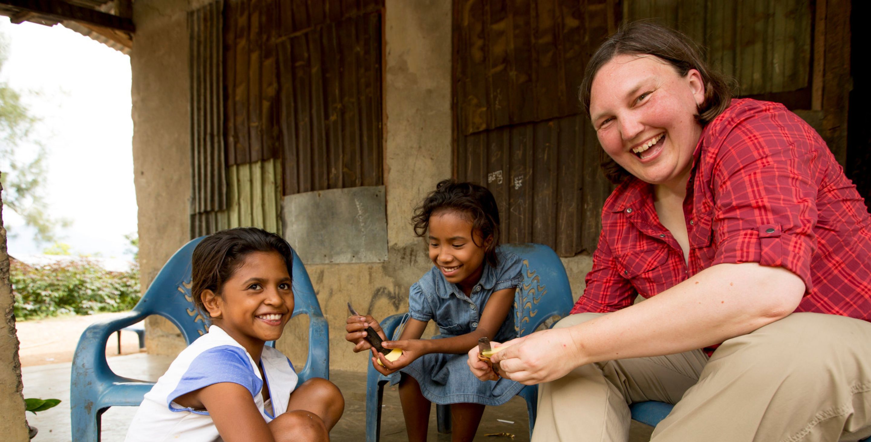An American woman sits with two little girls in Timor-Leste on their front porch, smiling as they enjoy a bowl of food.