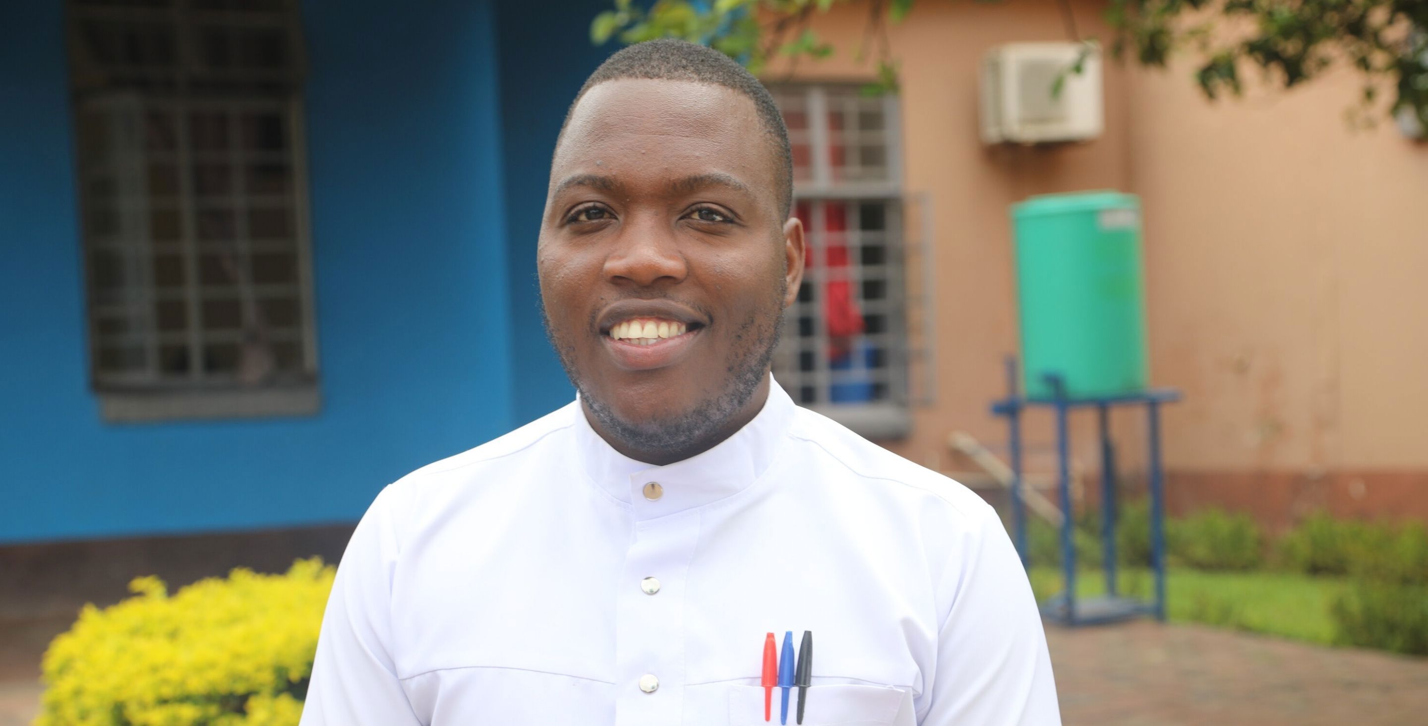 A young man in Zambia stands smiling at the camera in a nurses' uniform.