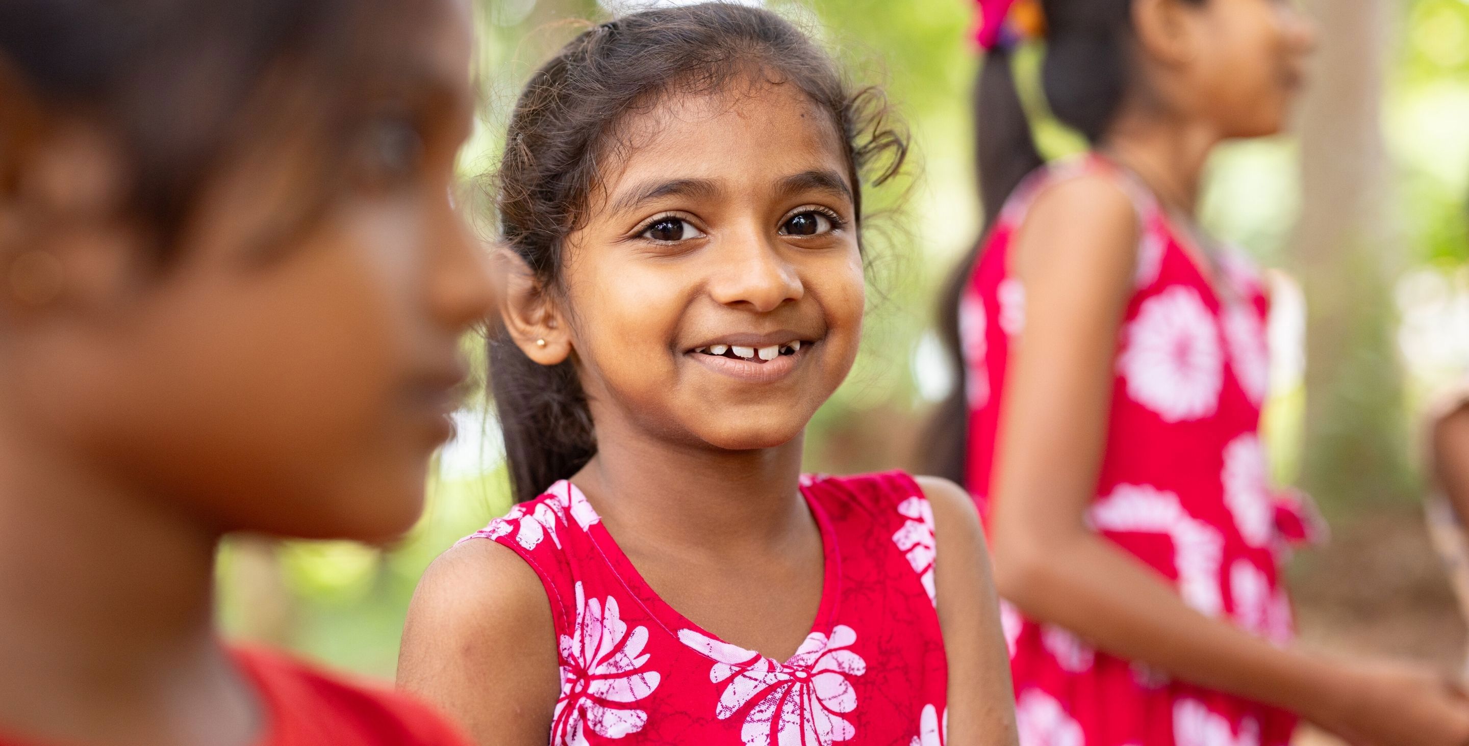 A girl wearing a red dress in Sri Lanka smiles outdoors while playing.