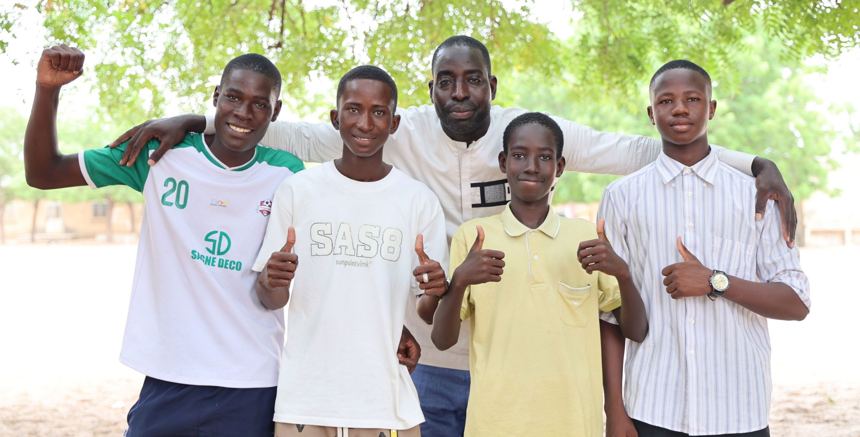 A group of men and young boys in Africa poses for a photo outdoors, some of them flexing their muscles, smiling happily.